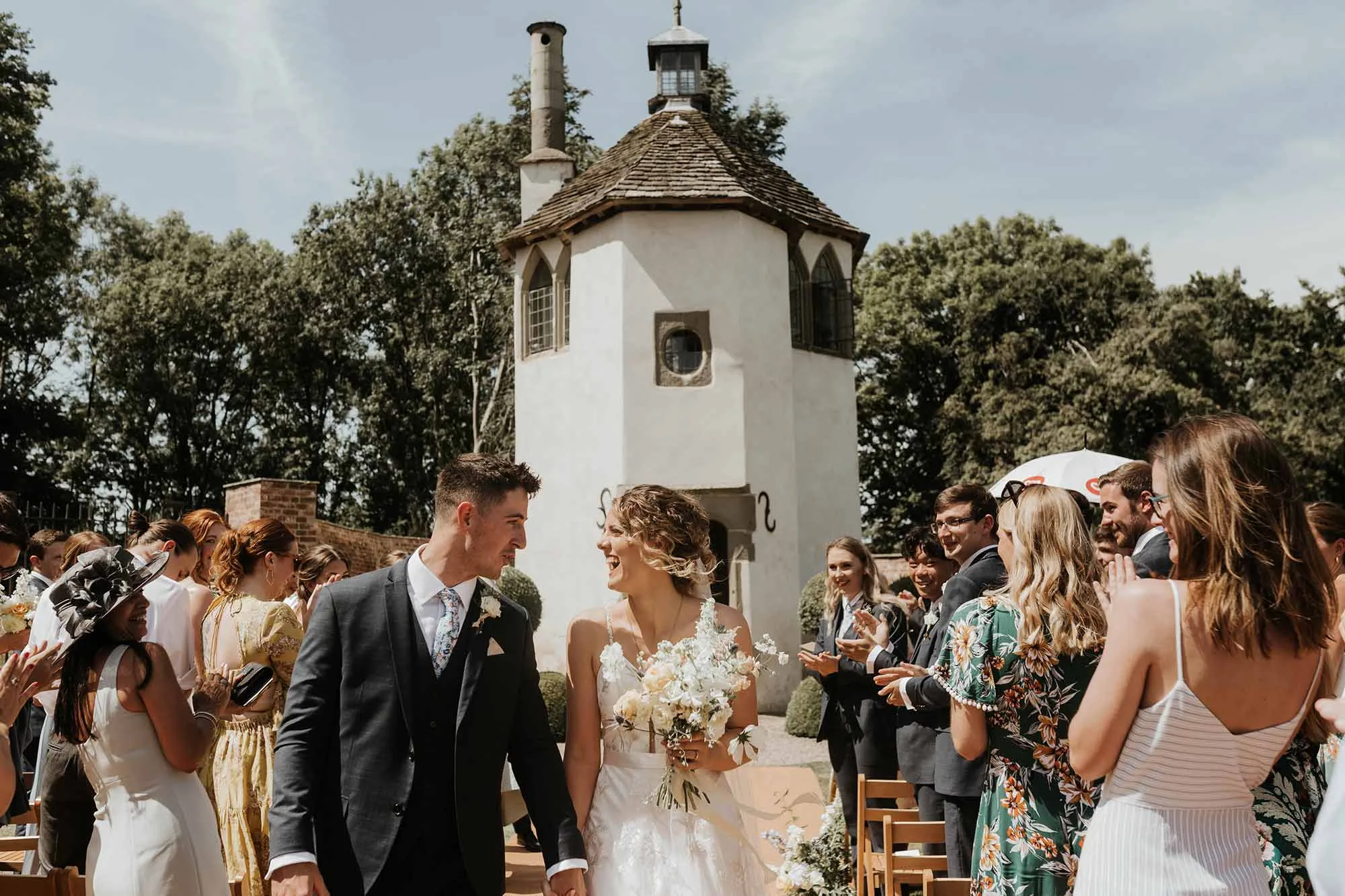 Bride-and-groom-walk-down-wedding-aisle-with-Homme-House-Summerhouse-in-the-background