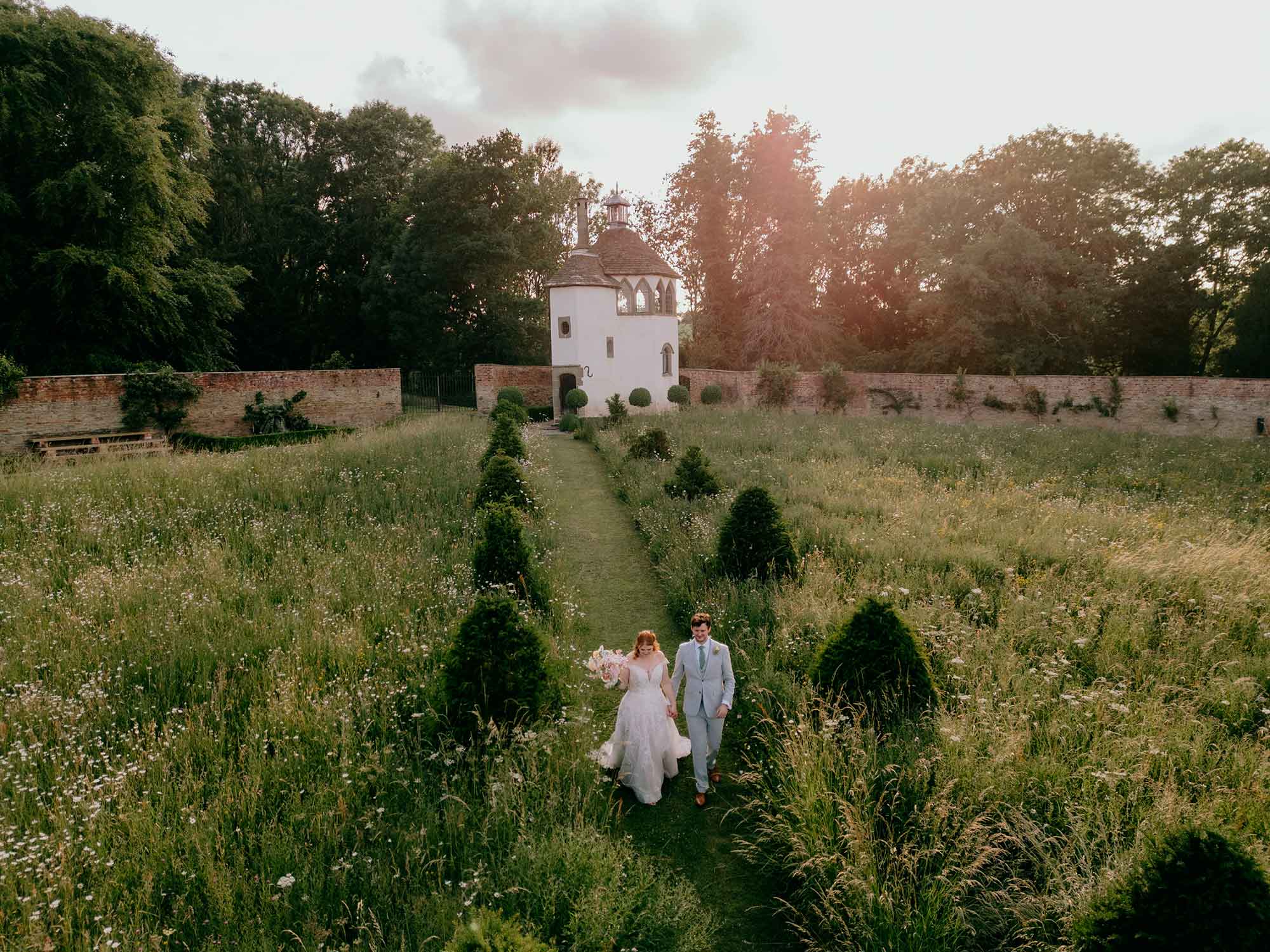 Bride-and-groom-walk-through-Homme-House-wild-flower-meadow-in-evening-sunshine