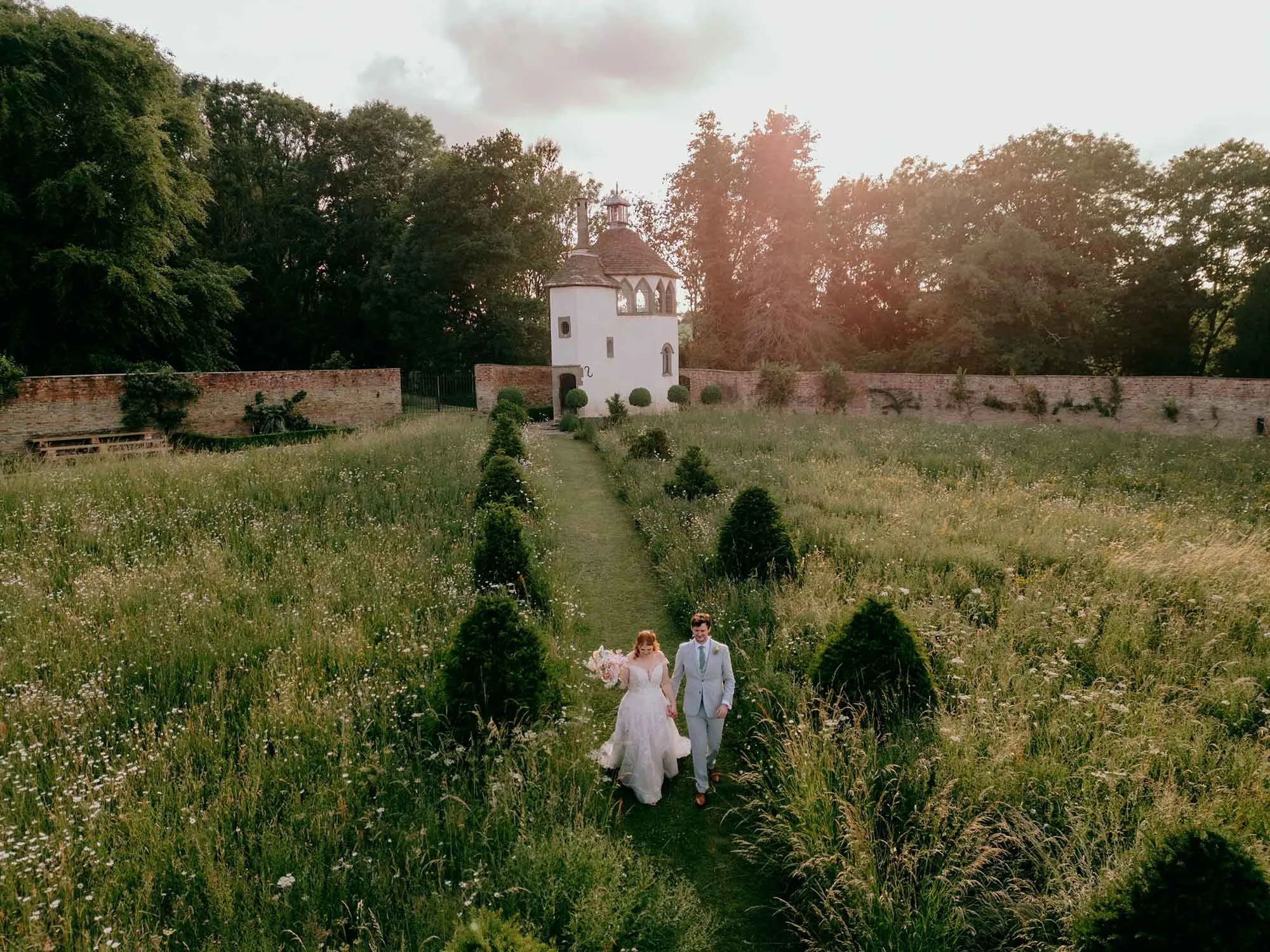 Bride-and-groom-walk-through-Homme-House-wild-flower-meadow-in-evening-sunshine