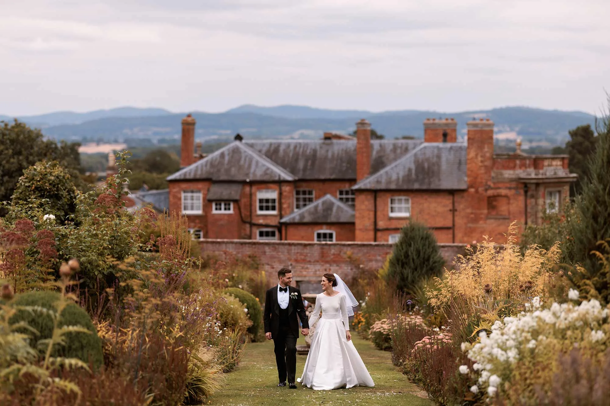 Bride-and-groom-walking-up-long-borders-in-walled-garden-at-Homme-House