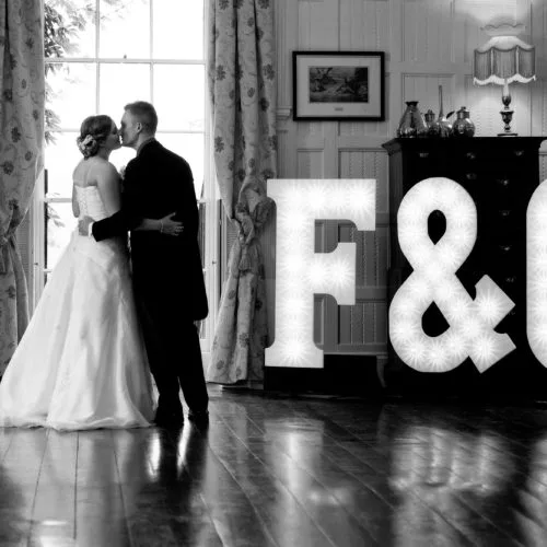 Bride and groom kiss beside light letters in the Panelled Room at Homme House