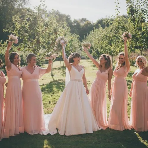 Bride and bridesmaids holding bouquets aloft in group photograph in walled garden orchard