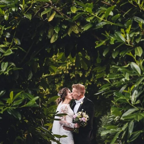 Bride and groom seen kissing through a laurel arch at Homme House