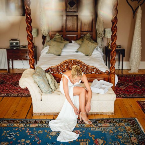 Bride-adjusts-shoe-on-chaise-longue-by-four-poster-bed
