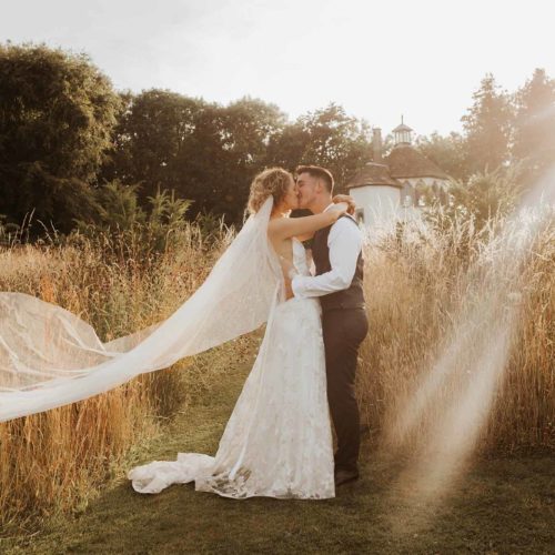 Bride-and-groom-kiss-with-floating-veil-in-front-of-Homme-House-wild-flower-meadow