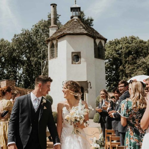 Bride-and-groom-walk-down-wedding-aisle-with-Homme-House-Summerhouse-in-the-background
